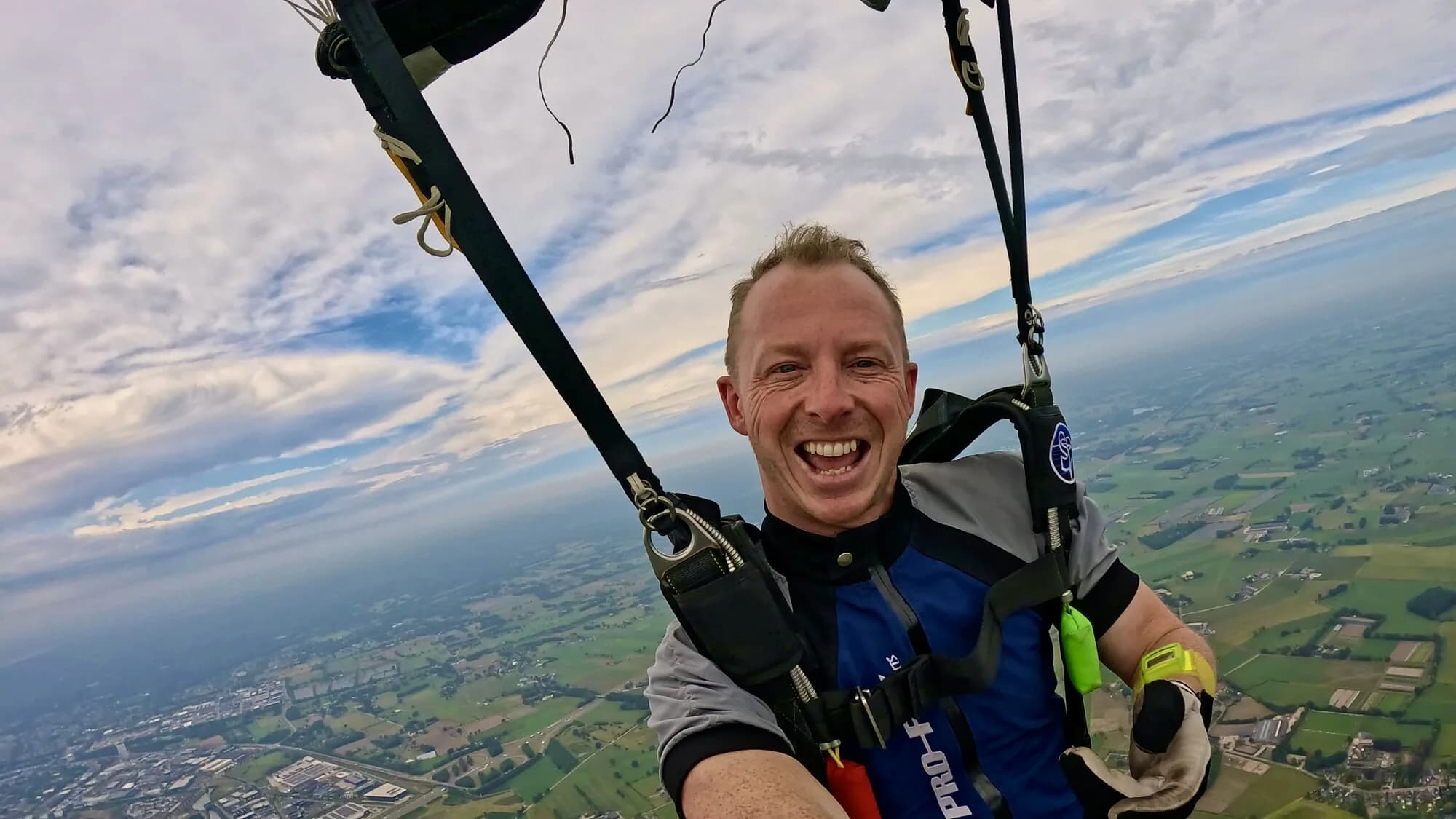Dennis lacht onder zijn parachute, hoog boven het landschap.